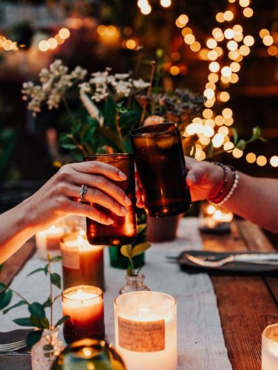 two people 'cheers' handmade glasses over a dinner table - covered in lit natural candles and foliage