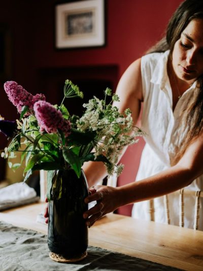Large vase made from a magnum champagne bottle with a bunch of flowers being placed on a table by a woman in a white dress