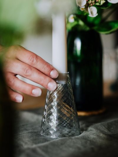 diamond glass candlestick holder with a cream tapered dinner candle being put on a table with a linen runner and repurposed glass vase in the background
